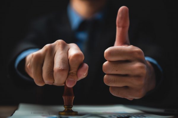 Close-up Of A Person's Hand Stamping With Approved Stamp On Document At Desk