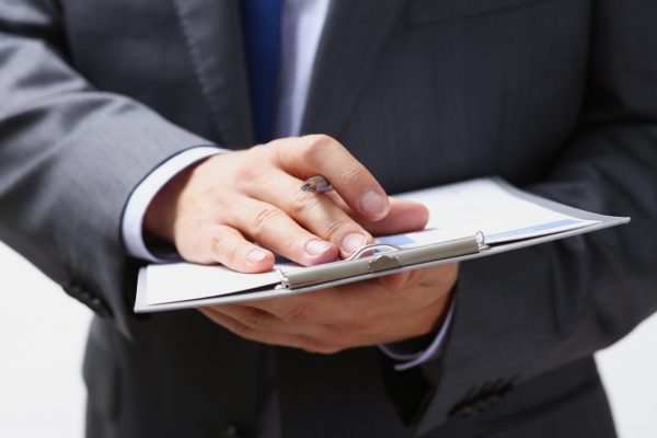 Male arm in suit and tie fill form clipped to pad with silver pen closeup. Sign gesture read pact sale agent bank job make note loan credit mortgage investment finance chief legal law concept