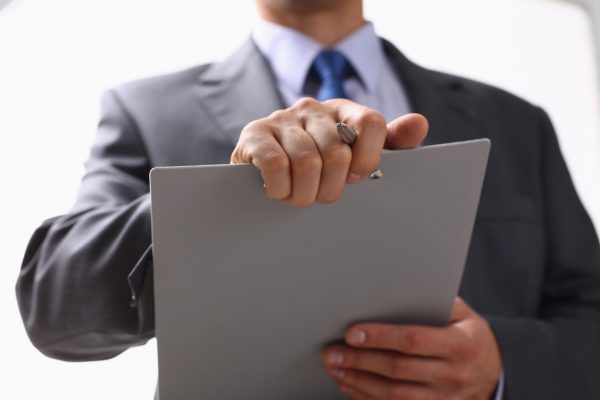 Hand of businessman in suit filling and signing with silver pen partnership agreement form clipped to pad closeup. Management training course, some important document, team leader ambition concept
